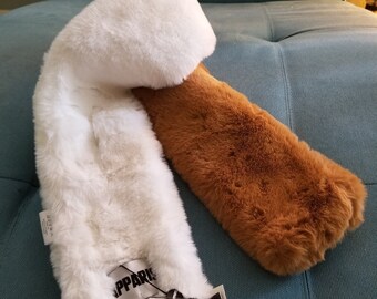 May include: A faux fur scarf in white and brown. The scarf is soft and fluffy, with a tag that reads "APPARI". The scarf is lying on a blue surface.