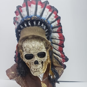 May include: A decorative skull bust wearing a headdress with red, white, and blue feathers. The skull is detailed with intricate carvings and has a gold finish.