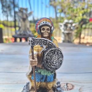 May include: A small, colorful figurine of a skeletal figure wearing a feathered headdress and holding a shield with a skull and crossbones design. The figure is standing on a black base.