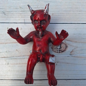 以下が含まれることがあります： 銀色の角と白い包帯を巻いた赤い悪魔の置物。置物は腕を伸ばして座っている。