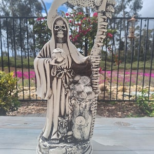May include: A white ceramic statue of a skeletal figure in a hooded robe, holding a skull in one hand and a bag of coins in the other. The figure stands on a base with a dragon's head and tail. An owl is perched at the base of the statue.