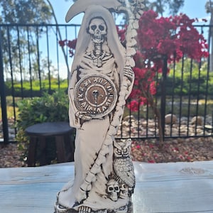 May include: A white ceramic statue of the Grim Reaper holding a scythe and standing on a pile of coins. The Reaper is wearing a hooded robe and has a skull face. An owl is perched on the Reaper's staff.