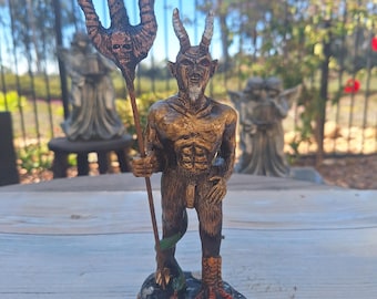 May include: A gold-colored figurine of a horned demon holding a trident. The demon has a beard and is standing on a black base.