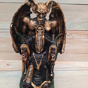 Puede incluir: Una figura esquelética negra y dorada se sienta en un trono con una criatura dorada con forma de murciélago detrás. El trono tiene acentos verdes y reposabrazos en forma de calavera.