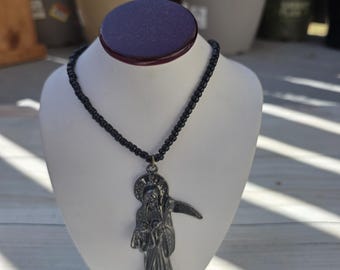 Puede incluir: Un collar con un colgante de metal oscuro con forma de figura encapuchada que sostiene una guadaña, suspendido de una cadena de pequeñas cuentas negras redondas. El colgante muestra detalles intrincados de la túnica y un halo circular detrás.