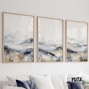 以下が含まれることがあります： 青、グレー、茶色の色合いで描かれた、霧のかかった山岳風景の水彩画が3枚、額装されています。絵画は、白とグレーのクッションのあるソファの上の白い壁に飾られています。