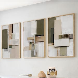 Puede incluir: Tres obras de arte abstractas enmarcadas con colores neutros. La obra presenta formas geométricas en tonos de blanco, beige y verde oliva. Los marcos son de madera clara. Un jarrón decorativo y otros objetos están en el estante de abajo.