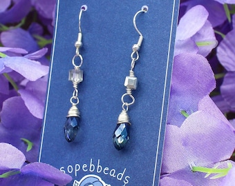 May include: A pair of silver dangle earrings with blue teardrop-shaped crystals and silver cube beads. The earrings are displayed on a blue card with the text "sopebeads HANDCRAFTED JEWELRY".