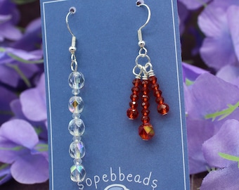 May include: A pair of earrings on a blue card with the text "sopebeads" and "HANDCRAFTED JEWELRY". One earring has a long strand of clear iridescent beads and the other earring has a cluster of red iridescent beads.