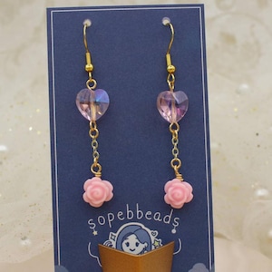 May include: Pink flower and iridescent heart dangle earrings with gold chain and hooks. The earrings are on a blue background with the text "sopebbeads".