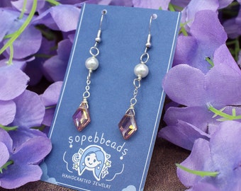 May include: A pair of handcrafted earrings with a silver-tone metal design. Each earring features a pearlescent bead, a faceted, diamond-shaped crystal with a color-shifting effect, and a fish hook closure. The earrings are displayed on a blue card with the text "sopebbeads".