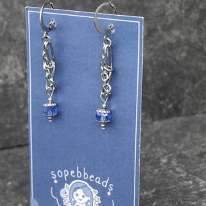 May include: A pair of silver chain earrings with blue crystal beads hanging from the bottom. The earrings are displayed on a blue card with the text "sopebbeads".