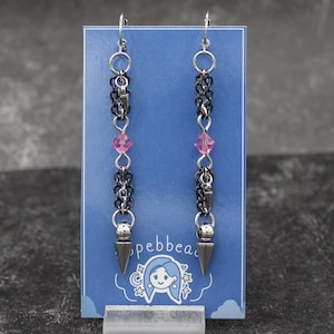 Puede incluir: Pendientes colgantes con ganchos plateados, eslabones de cadena negros y detalles de cristal rosa. Cada pendiente tiene un dije plateado en forma de cono en la parte inferior. Los pendientes se exhiben en una tarjeta azul con un logotipo.