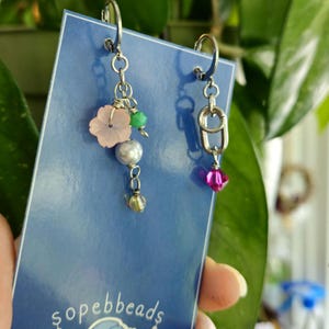 Puede incluir: Pendientes colgantes con ganchos plateados. Un pendiente presenta una flor rosa, cuentas verdes, una perla y una cuenta transparente. El otro pendiente tiene un cristal rosa. Los pendientes se exhiben en una tarjeta azul con el texto "sopebbeads".