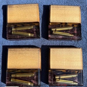 May include: Four square wooden coasters with a light brown top and a clear resin base. The resin contains several brass bullet casings. The coasters are arranged on a blue textured surface.