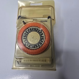 May include: A Sears brand electrical outlet timer in original plastic packaging. The device features a round, orange dial with black and white markings for AM and PM hours, and a central gold-colored face with the word TIME printed on it.