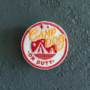 May include: Circular embroidered patch with a white border and a red and yellow design. The patch reads "CAMP DOG" in yellow script, with "ON DUTY" in red. A red mountain and tent graphic is in the centre.