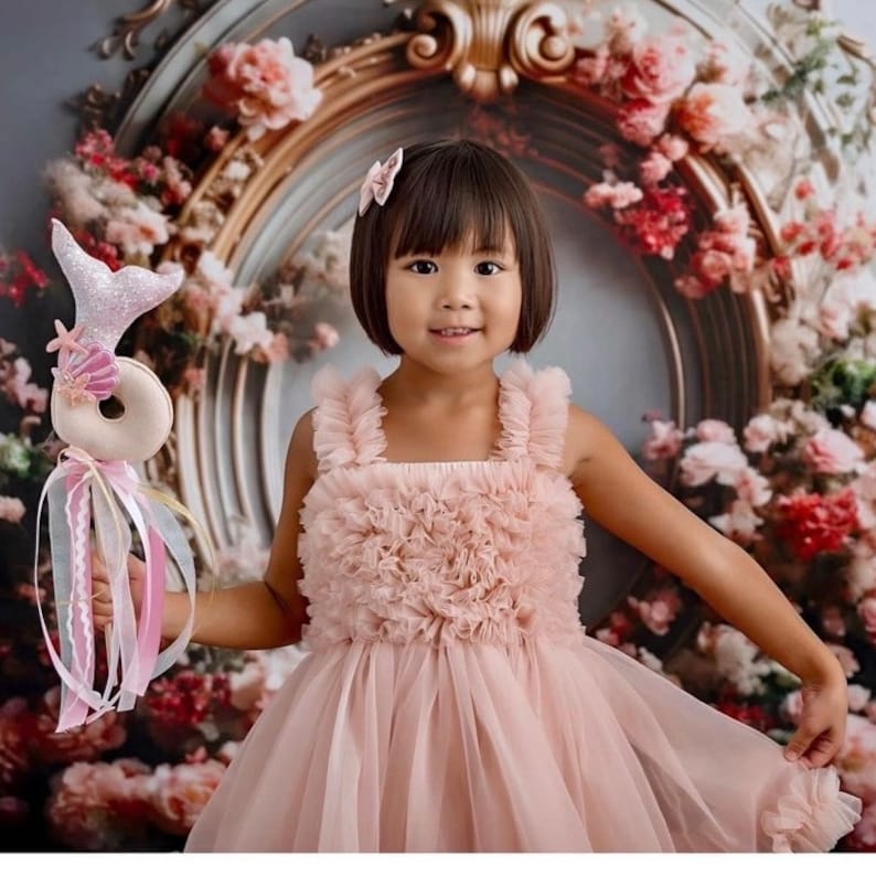 May include: A young girl wearing a pink tulle dress with ruffled detailing holds a pink and silver mermaid wand with a donut-shaped charm. The background is a floral backdrop with a gold ornate frame.