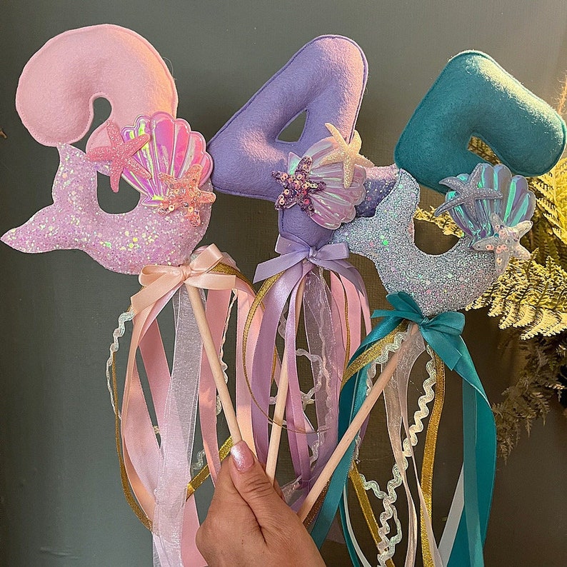 May include: Three mermaid-themed wands with numbers 1, 4, and 5. Each wand has a glittery mermaid tail, a felt number, and a seashell with a starfish. The wands are decorated with pink, purple, and teal ribbons.