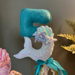 May include: A turquoise felt number five with a glittery mermaid tail and iridescent seashells. This mermaid-themed topper is perfect for a birthday cake or party decoration.