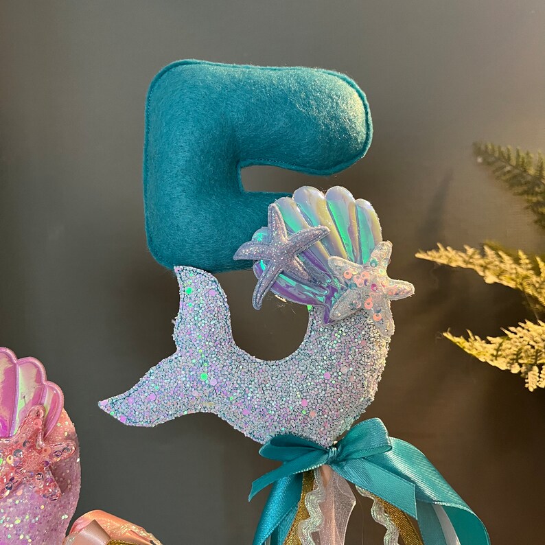 May include: A turquoise felt number five with a glittery mermaid tail and iridescent seashells. This mermaid-themed topper is perfect for a birthday cake or party decoration.