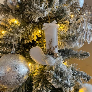 Peut inclure: Une décoration de Noël en forme de chaussette blanche avec un nœud, des détails floraux et une perle dorée est suspendue à un sapin de Noël enneigé. Une boule de Noël argentée et d'autres décorations sont également visibles.