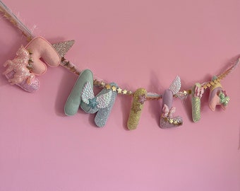 May include: A pastel-colored felt letter banner with the word "MIA" spelled out. Each letter is decorated with glitter, stars, and wings. The banner is strung on a gold string with white feathers.