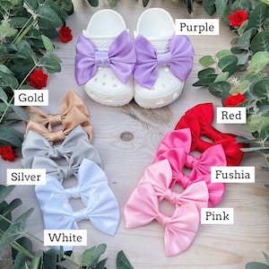 May include: A set of ten satin bows in various colours, including purple, gold, silver, white, red, fuchsia, and pink. The bows are arranged in two rows on a wooden surface.