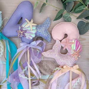 May include: Two purple, pink, and blue felt mermaid wands with glitter and seashell accents. The wands are decorated with ribbons and are perfect for a mermaid-themed party.