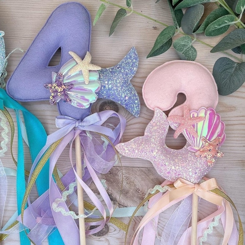 May include: Two purple, pink, and blue felt mermaid wands with glitter and seashell accents. The wands are decorated with ribbons and are perfect for a mermaid-themed party.