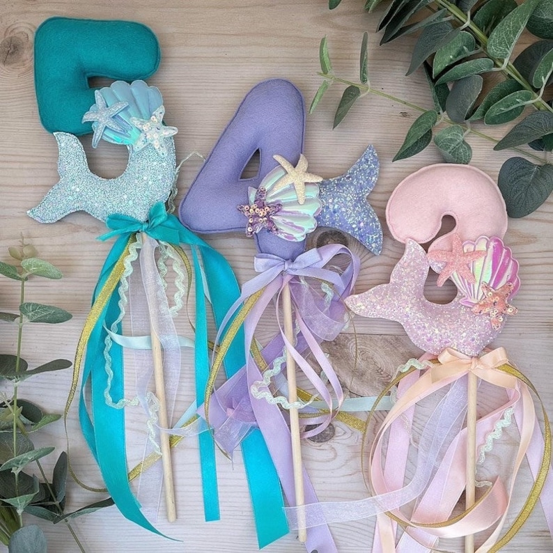 May include: Three mermaid-themed wands with glitter and felt numbers 5, 4, and 3. Each wand has a glittery mermaid tail, a seashell, and a starfish. The wands are decorated with blue, purple, and pink ribbons.
