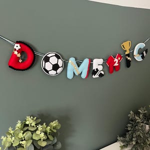 May include: A felt banner with the name "DOMENIC" spelled out with individual letters and soccer-themed embellishments. The banner includes a soccer ball, a trophy, and a jersey with the number 2. The letters are in various colors.