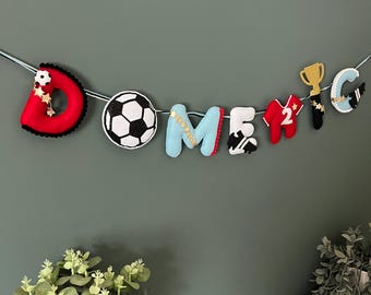 Puede incluir: Una guirnalda de fieltro con el nombre "DOMENIC" escrito con letras individuales y adornos temáticos de fútbol. La guirnalda incluye un balón de fútbol, un trofeo y una camiseta con el número 2. Las letras son de varios colores.