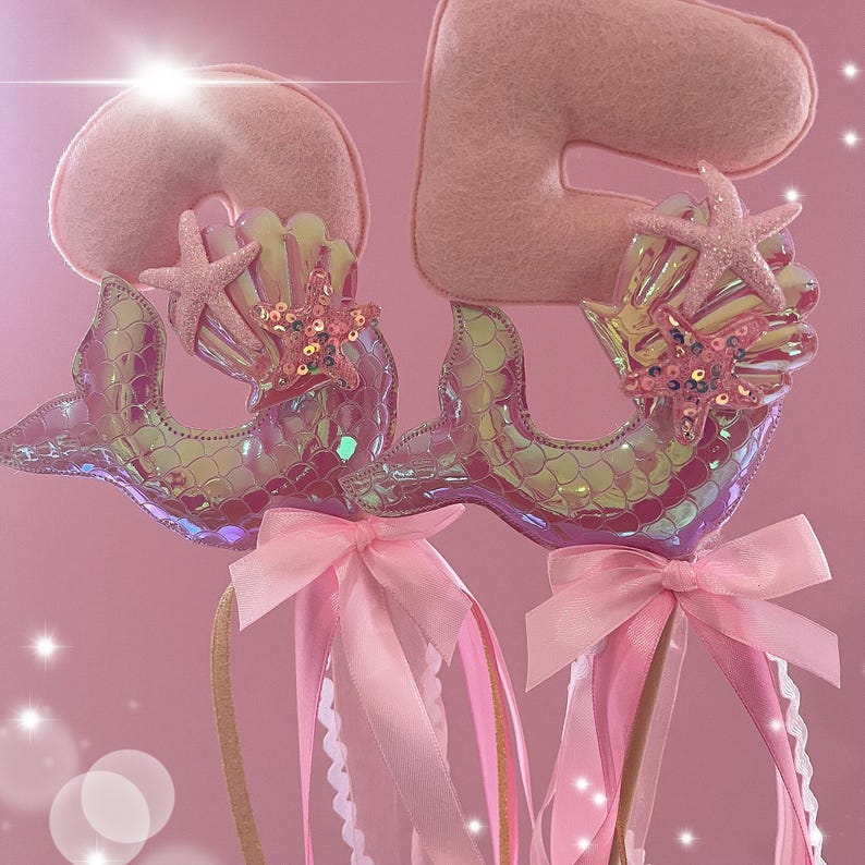 May include: Two pink felt mermaid tail wands with iridescent scales, pink bows, and glitter seashells. Each wand has a pink felt number, one is a 2 and the other is a 5.