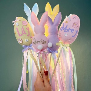 May include: Four pastel-coloured Easter bunny wands with felt bunny heads and egg-shaped decorations. The eggs are decorated with glitter and the names "Lily", "Aurelia", and "Aubrey".