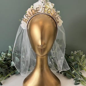 May include: White bridal headband with a veil and a flower crown. The headband has the words "Bride To Be" in rhinestones.