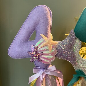 May include: Purple felt number four with iridescent seashells and glitter starfish decorations. The number is attached to a purple ribbon and a stick.