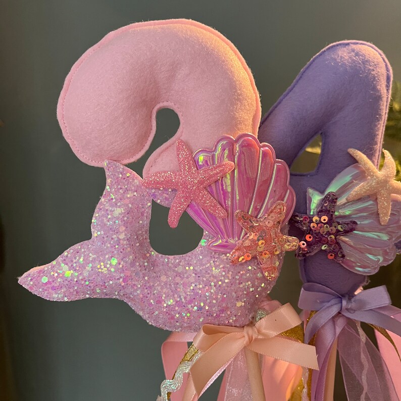 May include: Two purple and pink felt mermaid tail wands with glitter and iridescent seashells. The wands are decorated with pink and purple ribbons.