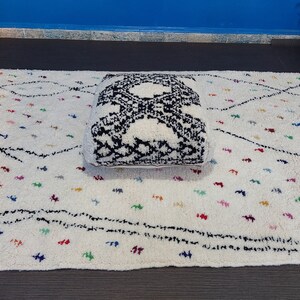 Puede incluir: Un coj&iacute;n de suelo blanco y negro con flecos sobre una alfombra blanca con l&iacute;neas negras y puntos de colores.