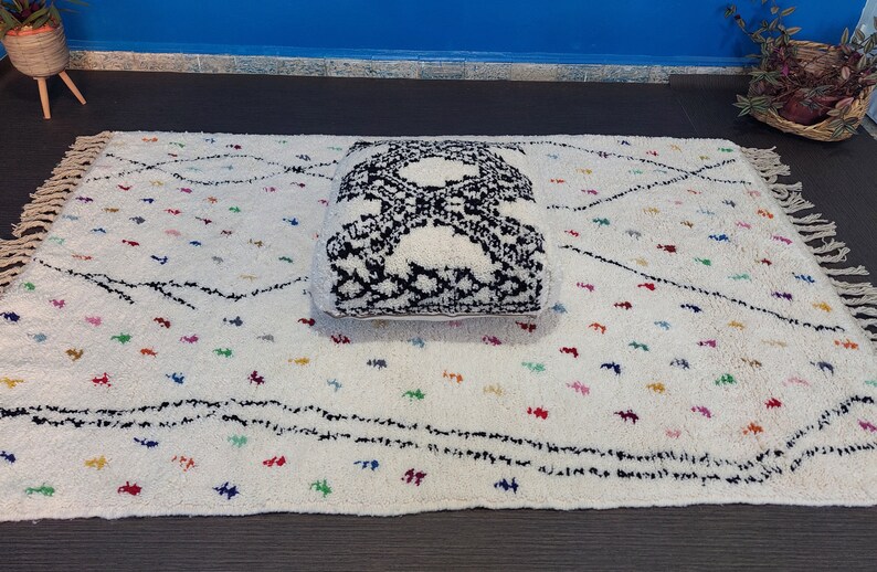 Puede incluir: Un coj&iacute;n de suelo blanco y negro con flecos sobre una alfombra blanca con l&iacute;neas negras y puntos de colores.