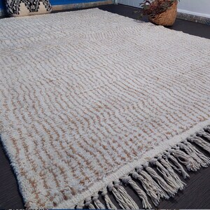 May include: A large, rectangular rug with a cream-colored background and a textured, wavy pattern in light brown. The rug has a fringe along one edge. The rug is placed on a dark gray surface.