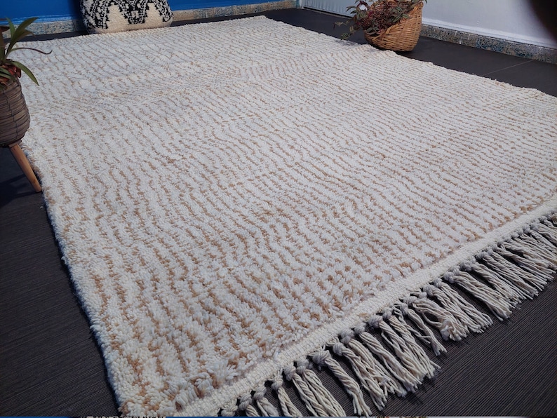 May include: A large, rectangular rug with a cream-colored background and a textured, wavy pattern in light brown. The rug has a fringe along one edge. The rug is placed on a dark gray surface.