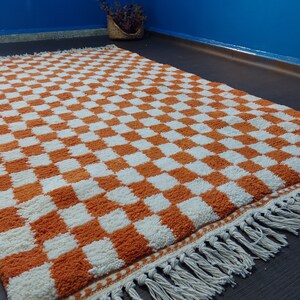 Op de afbeelding: Een rechthoekig vloerkleed met een oranje en wit dambordpatroon. Het kleed heeft een dikke pool en een franjerand. Gemaakt van zacht materiaal, geschikt voor een woon- of slaapkamer. De afmetingen van het kleed zijn in centimeters.