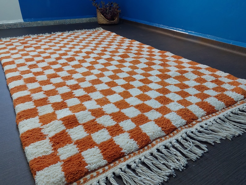 Op de afbeelding: Een rechthoekig vloerkleed met een oranje en wit dambordpatroon. Het kleed heeft een dikke pool en een franjerand. Gemaakt van zacht materiaal, geschikt voor een woon- of slaapkamer. De afmetingen van het kleed zijn in centimeters.