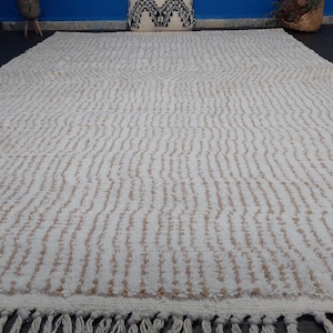 May include: Cream-colored, textured rug with a repeating vertical pattern and a fringe along the bottom edge. The rug has a central design element and is suitable for home decor.