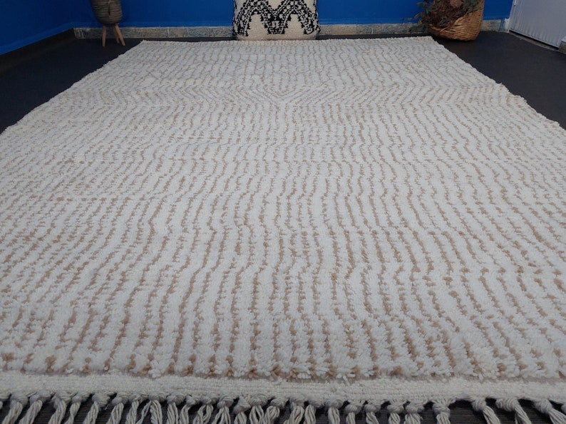 May include: Cream-colored, textured rug with a repeating vertical pattern and a fringe along the bottom edge. The rug has a central design element and is suitable for home decor.