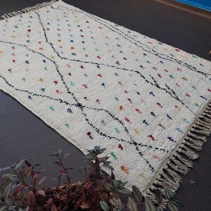 Puede incluir: Una alfombra blanca con un patr&oacute;n geom&eacute;trico negro y acentos de colores. La alfombra tiene flecos en todos los lados.
