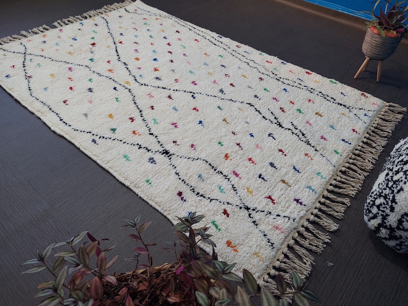 Puede incluir: Una alfombra blanca con un patr&oacute;n geom&eacute;trico negro y acentos de colores. La alfombra tiene flecos en todos los lados.
