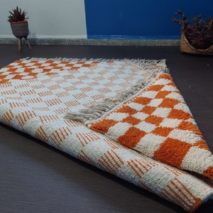 Op de afbeelding: Een opgerolde, rechthoekige vloerkleed met een herhalend oranje en wit geblokt patroon. Het vloerkleed heeft een franje langs &eacute;&eacute;n rand. Het patroon is zichtbaar aan beide zijden van het opgerolde gedeelte. Het vloerkleed is gemaakt van een textuurrijk materiaal.