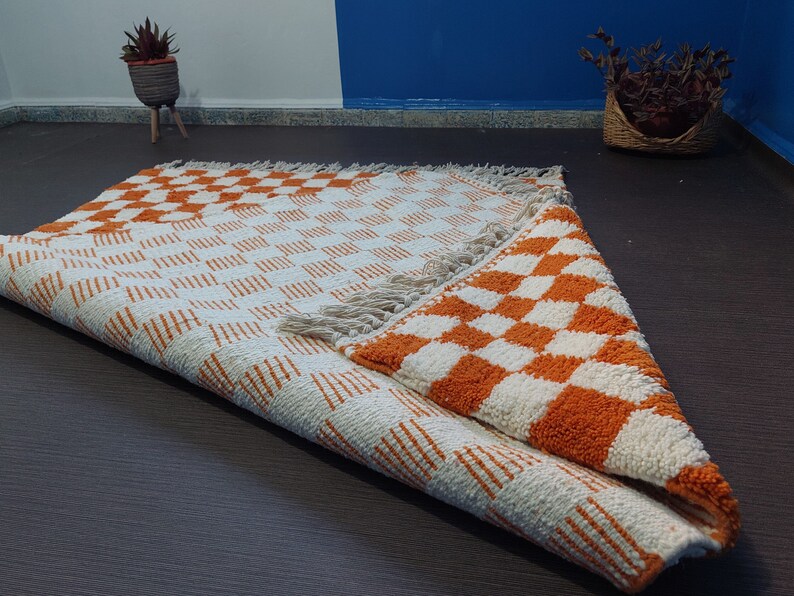 Op de afbeelding: Een opgerolde, rechthoekige vloerkleed met een herhalend oranje en wit geblokt patroon. Het vloerkleed heeft een franje langs &eacute;&eacute;n rand. Het patroon is zichtbaar aan beide zijden van het opgerolde gedeelte. Het vloerkleed is gemaakt van een textuurrijk materiaal.